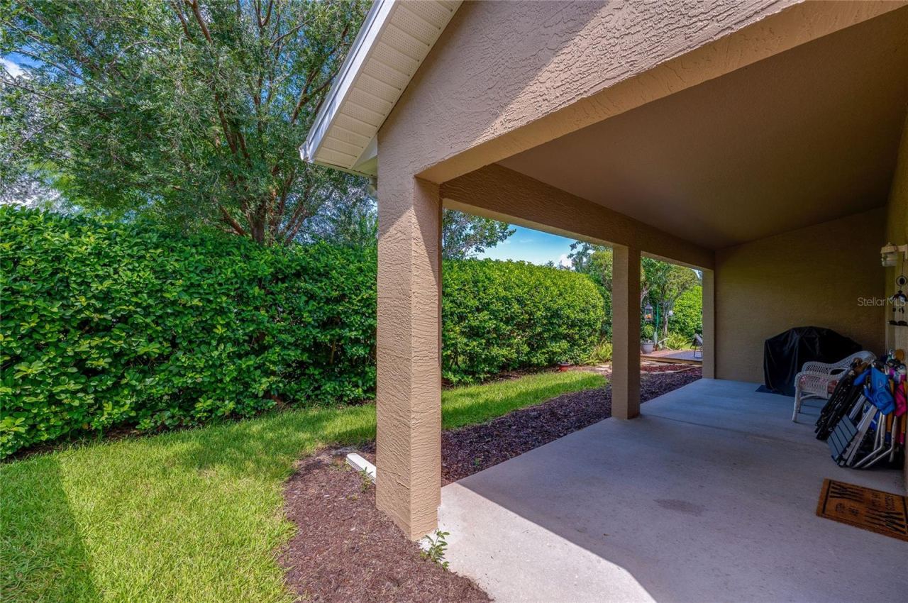 Photo 20 of 20: Covered concrete back patio offering protection from the elements alongside a manicured green lawn. Tall, dense privacy hedges frame the perimeter to create a quiet outdoor sanctuary.