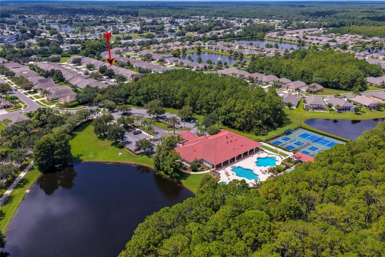 Photo 3 of 20: Elevated wide-angle aerial perspective highlighting the community clubhouse, sports courts, and adjacent lake, with an arrow pointing toward the neighborhood homes nestled behind green tree lines.
