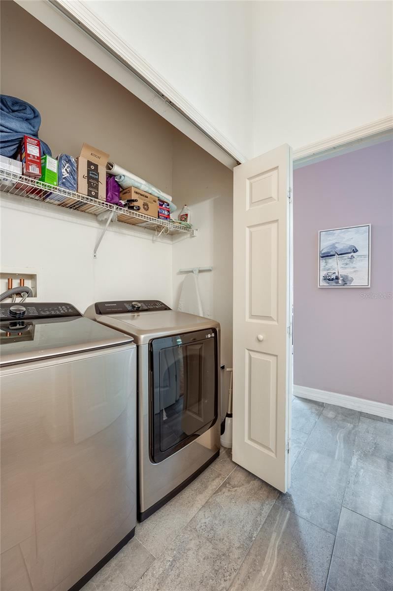Photo 17 of 20: Dedicated laundry alcove equipped with side-by-side stainless steel appliances and overhead wire storage shelving. Grey tile flooring connects the utility space to the adjacent living areas.