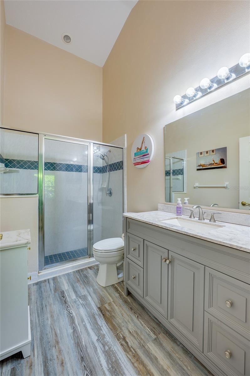 Photo 13 of 20: En-suite bathroom featuring a grey vanity with marble counter, modern laminate flooring, and vanity lighting. A glass-enclosed walk-in shower with decorative blue accent tile completes the space.