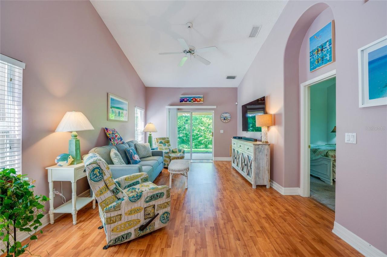 Photo 9 of 20: Spacious living room boasting dramatic vaulted ceilings, a central ceiling fan, and warm laminate flooring. Large sliding glass doors open directly to the outdoor patio space, while an arched doorway provides access to an adjoining bedroom.