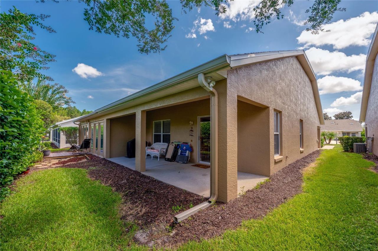 Photo 19 of 20: Exterior view of the rear elevation featuring a long covered patio and a well-maintained lawn bordered by mulch beds. Crisp stucco finishes and integrated roof gutters complete the exterior structure under clear blue skies.