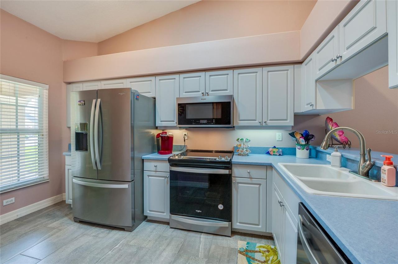 Photo 5 of 20: Functional kitchen equipped with stainless steel appliances, white cabinetry, and blue countertops. A large window brings in natural light, while high vaulted ceilings add depth.