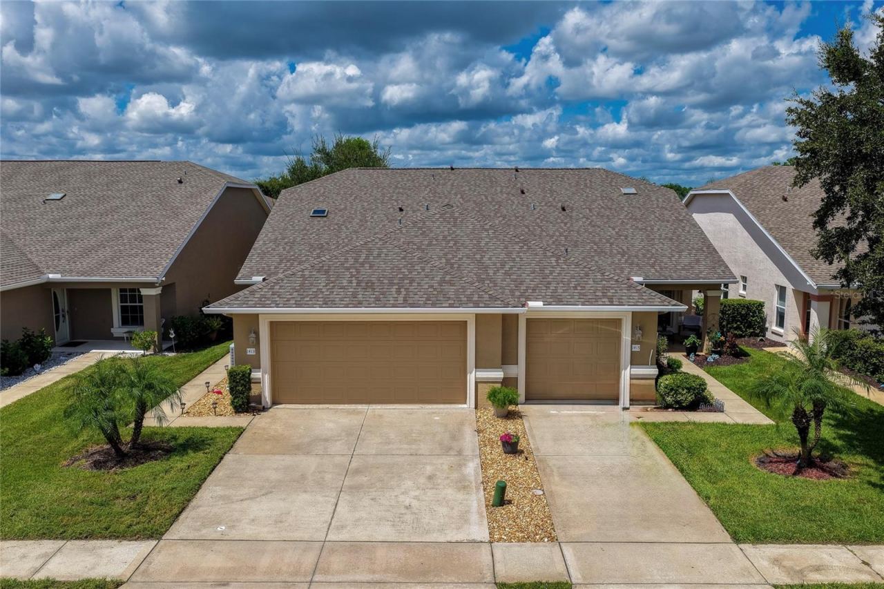 Photo 2 of 20: Front-facing view of the home featuring neutral stucco siding, an attached garage, and an expansive paved driveway surrounded by tropical green landscaping.