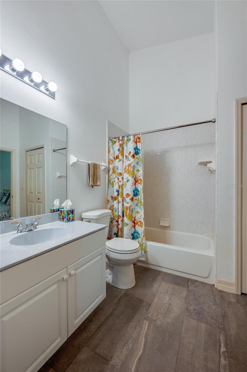 Photo 16 of 20: Functional bathroom featuring dark tile flooring, a wide vanity mirror, and bright overhead lighting. The combination shower and bathtub includes built-in wall shelving and tiled surrounds.