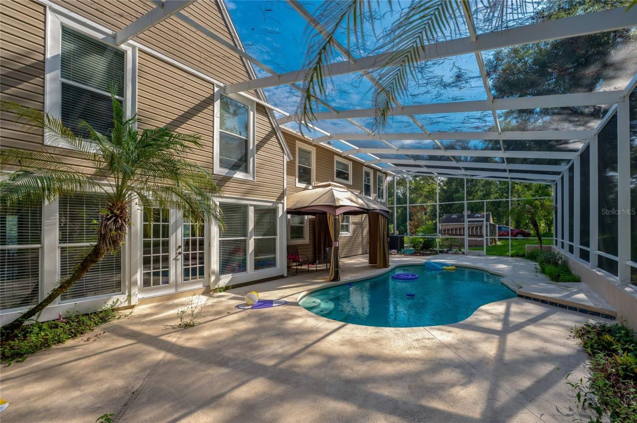 Photo 2 of 20: Sunlit outdoor pool deck enclosed in a screened lanai, highlighting French doors leading inside and lush tropical landscaping.