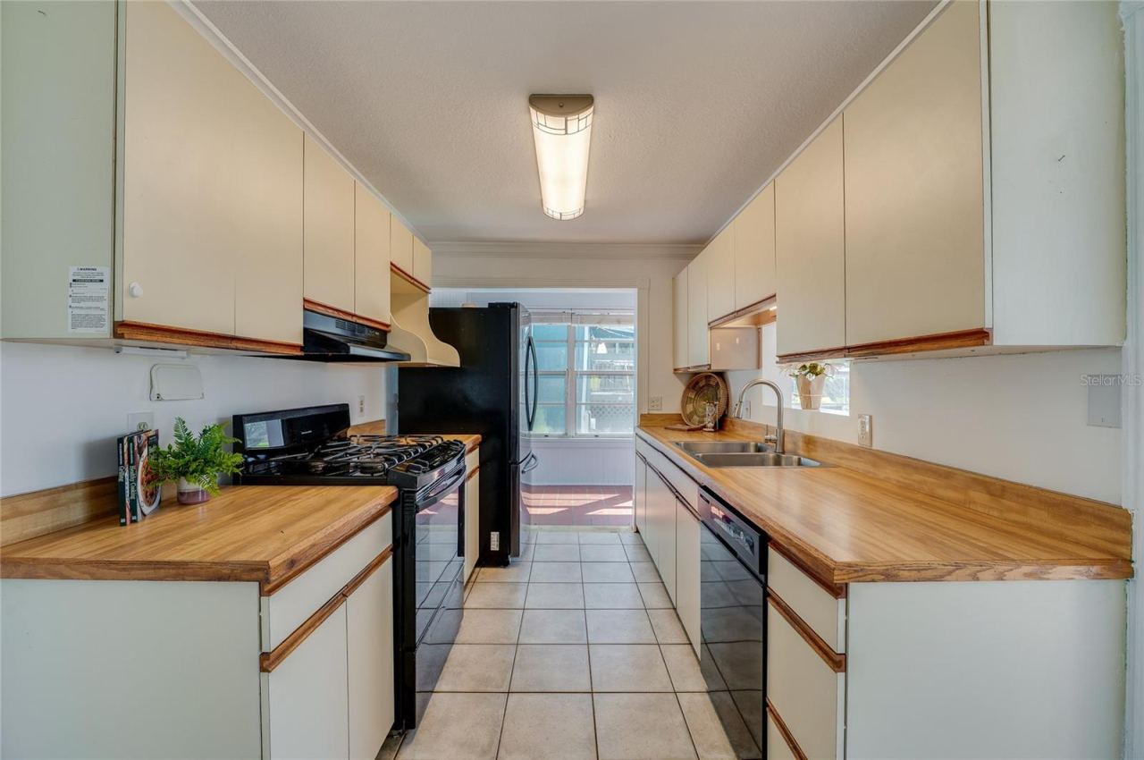 Photo 10 of 20: Galley-style kitchen design equipped with dual-sided cabinetry, warm wood-toned countertops, a double-basin sink, and overhead light fixtures. The walkthrough layout opens up directly to the sunroom beyond.