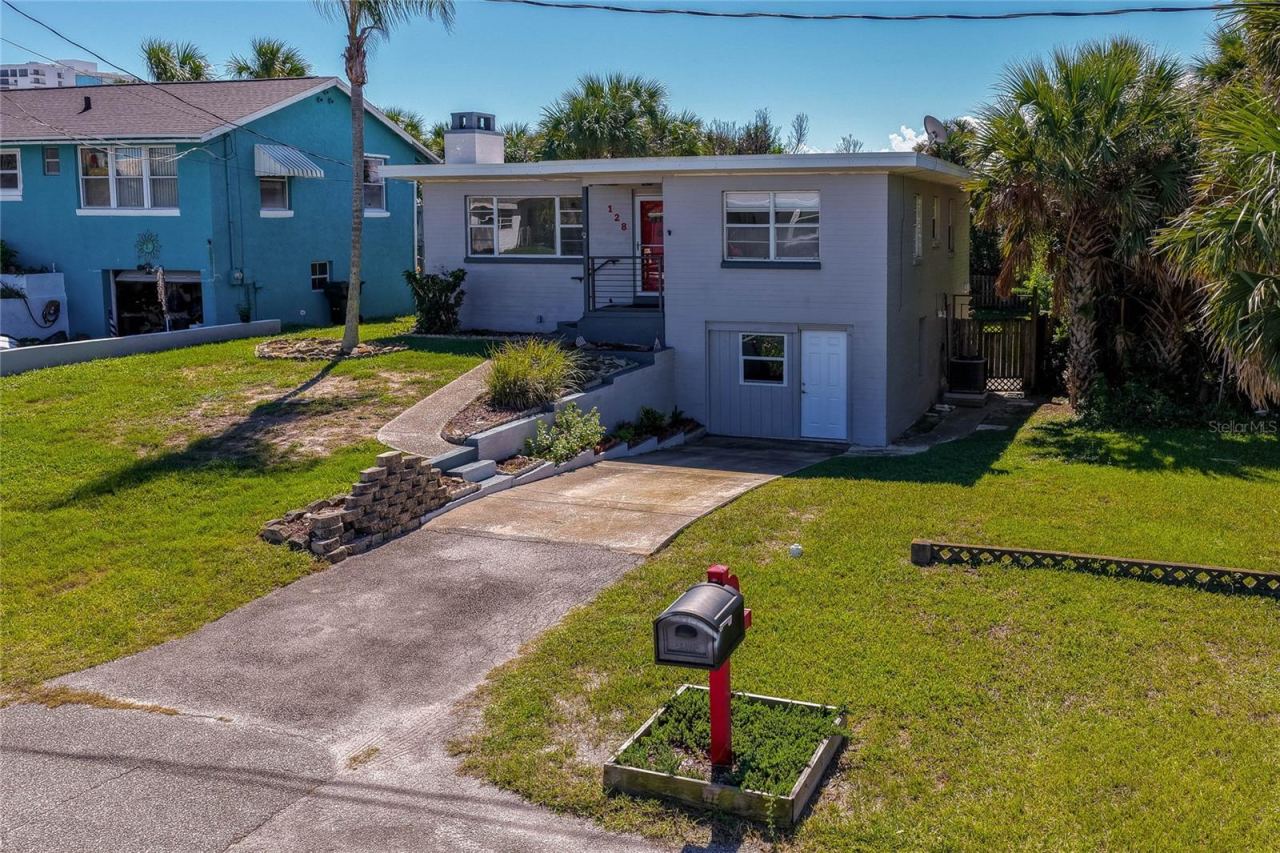 Photo 4 of 20: Front exterior perspective of the two-story residence showcasing a paved driveway, a red mailbox fixture, and mature palm trees. The sloped front yard is framed by stone retaining wall accents.