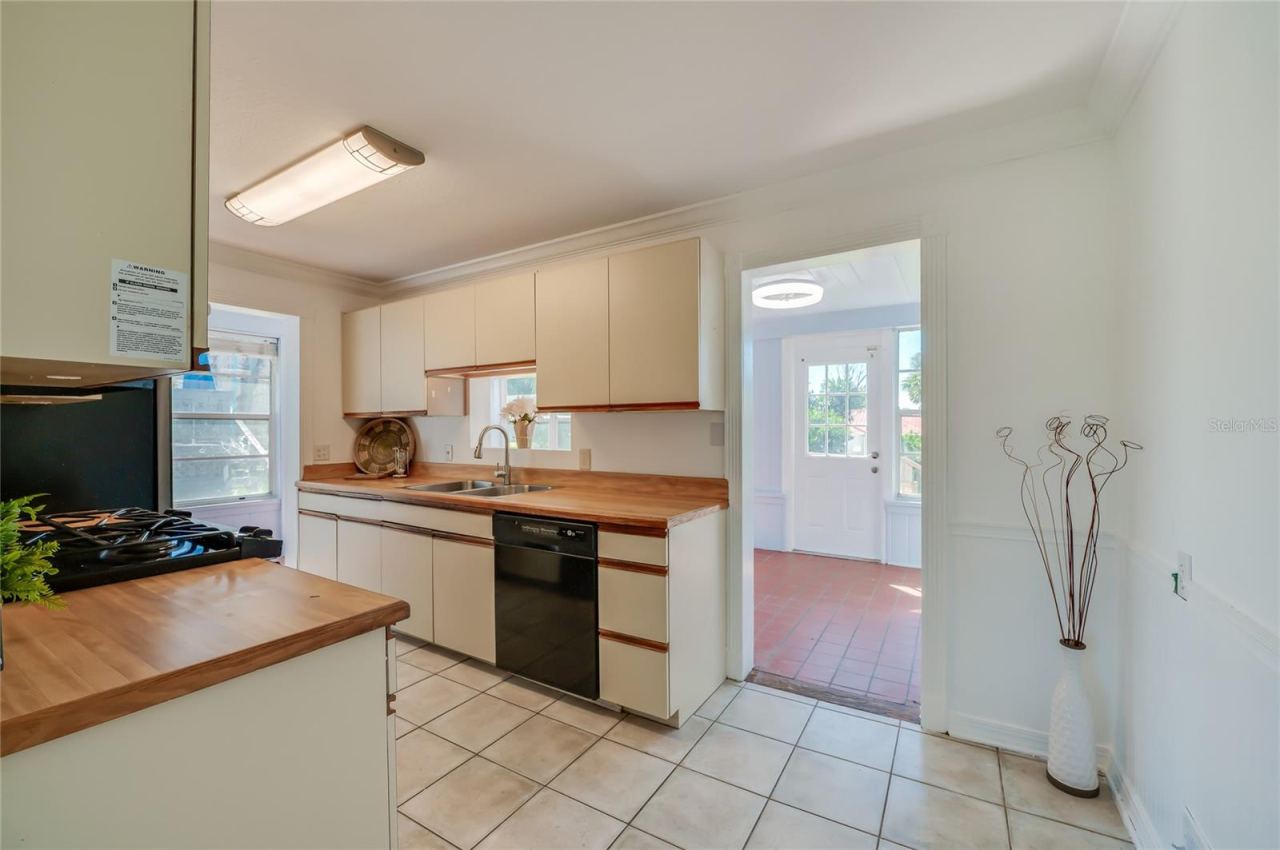 Photo 8 of 20: Functional kitchen featuring butcher block-style countertops, classic cabinetry, tile flooring, and integrated black appliances. An adjacent open doorway provides direct access to a sunlit room with terracotta tile floors.