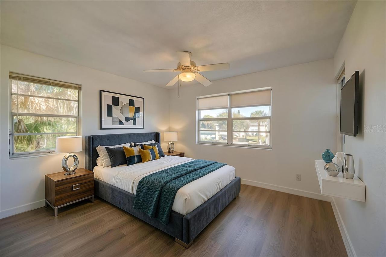 Photo 18 of 20: Virtually Staged - Bright bedroom area framed by continuous LVP flooring, smooth white walls, and a traditional white ceiling fan. Dual aspect windows bring in abundant sunlight and views of the surrounding palm trees.