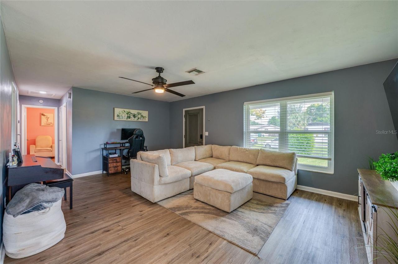 Photo 7 of 20: Generously sized main room featuring LVP flooring, soft gray walls, a central ceiling fan, and large front-facing windows.