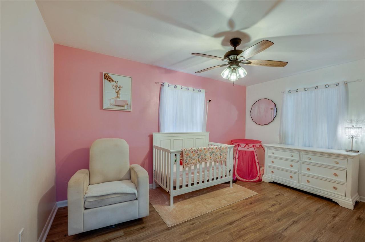Photo 17 of 20: Bright bedroom area highlighted by warm LVP flooring, soft pink accent walls, a ceiling fan, and ample natural light from two windows.