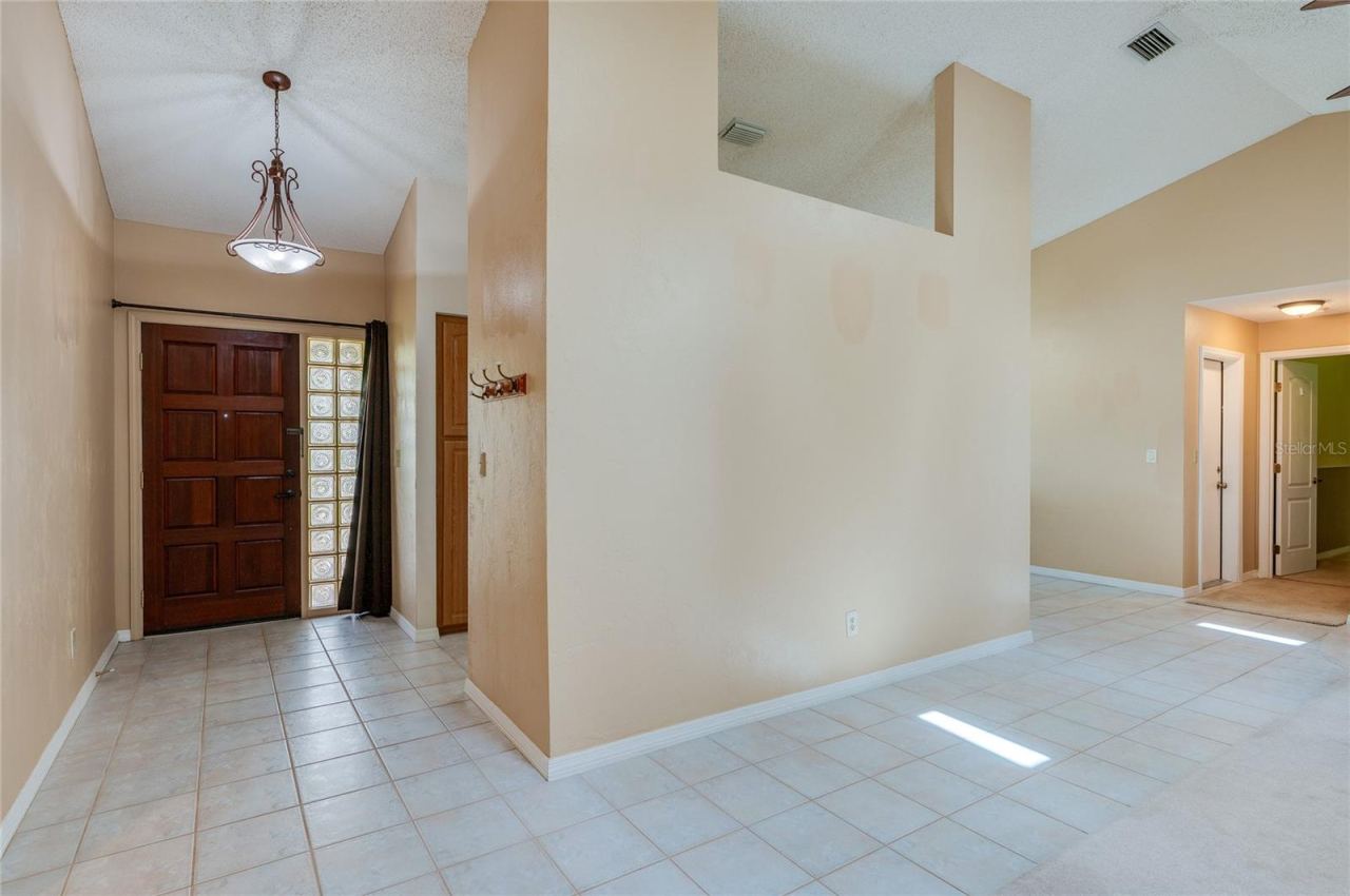 Photo 6 of 20: Spacious entry hallway featuring tile flooring, warm neutral walls, and a large decorative wood door with glass block side panels. Vaulted ceilings and open wall cutouts create a light-filled ambiance.